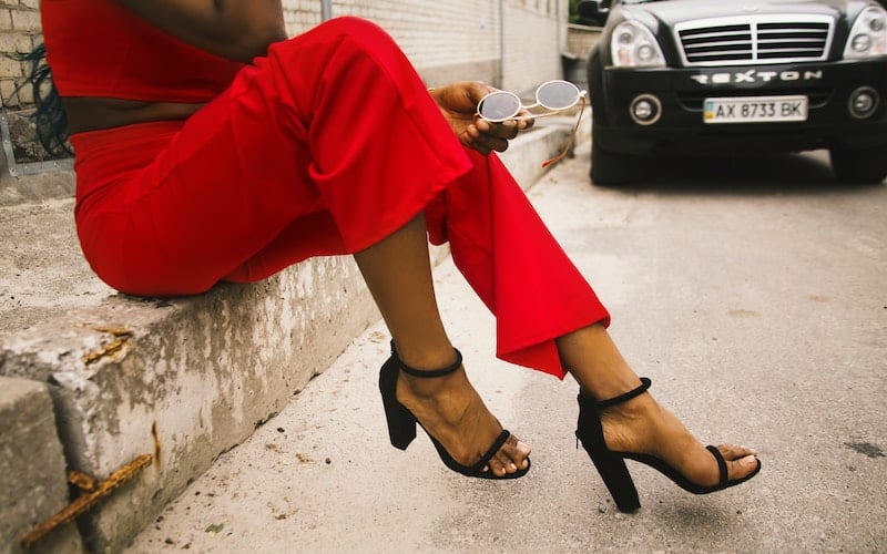 a woman wearing a red pant suit and black heels sitting on the curb